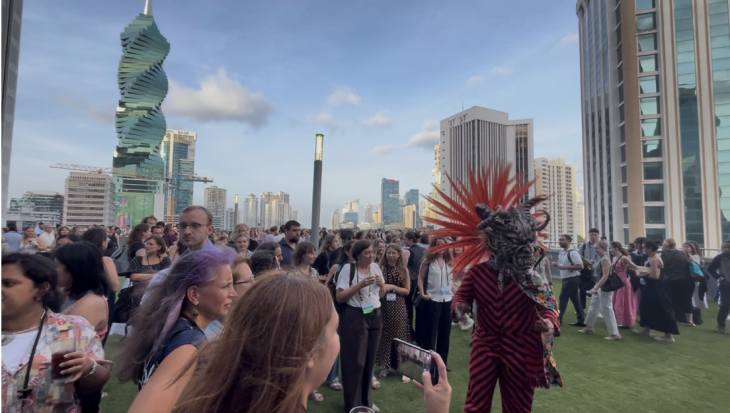 A crowd gathers on a rooftop overlooking a city skyline as a performer in an elaborate red feathered headdress and patterned costume interacts with attendees. Several people watch and take photos during the outdoor event at sunset.